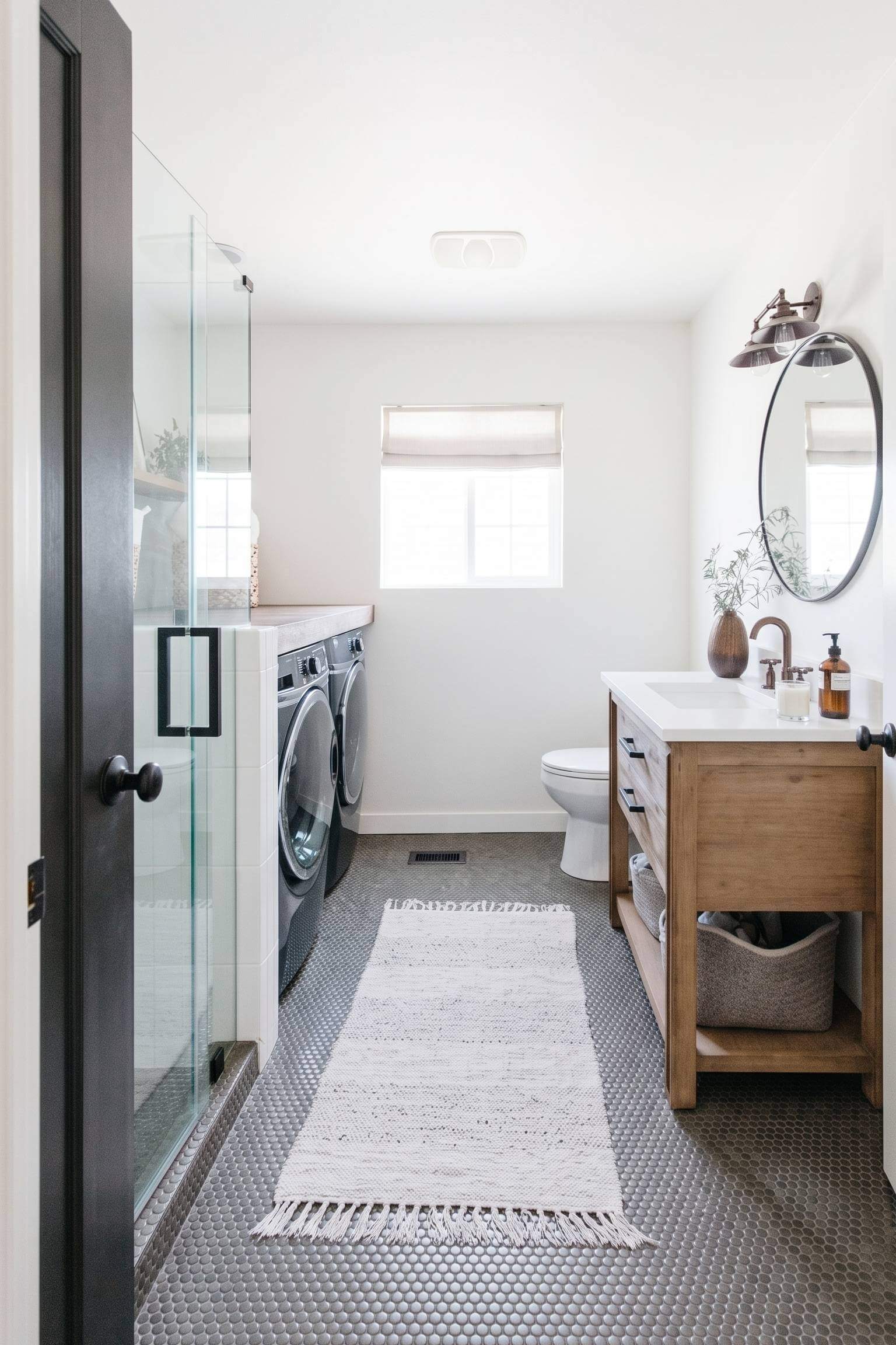A dramatic bathroom featuring a bold black interior door, gray penny tiles, and laundry machines tucked by the shower.